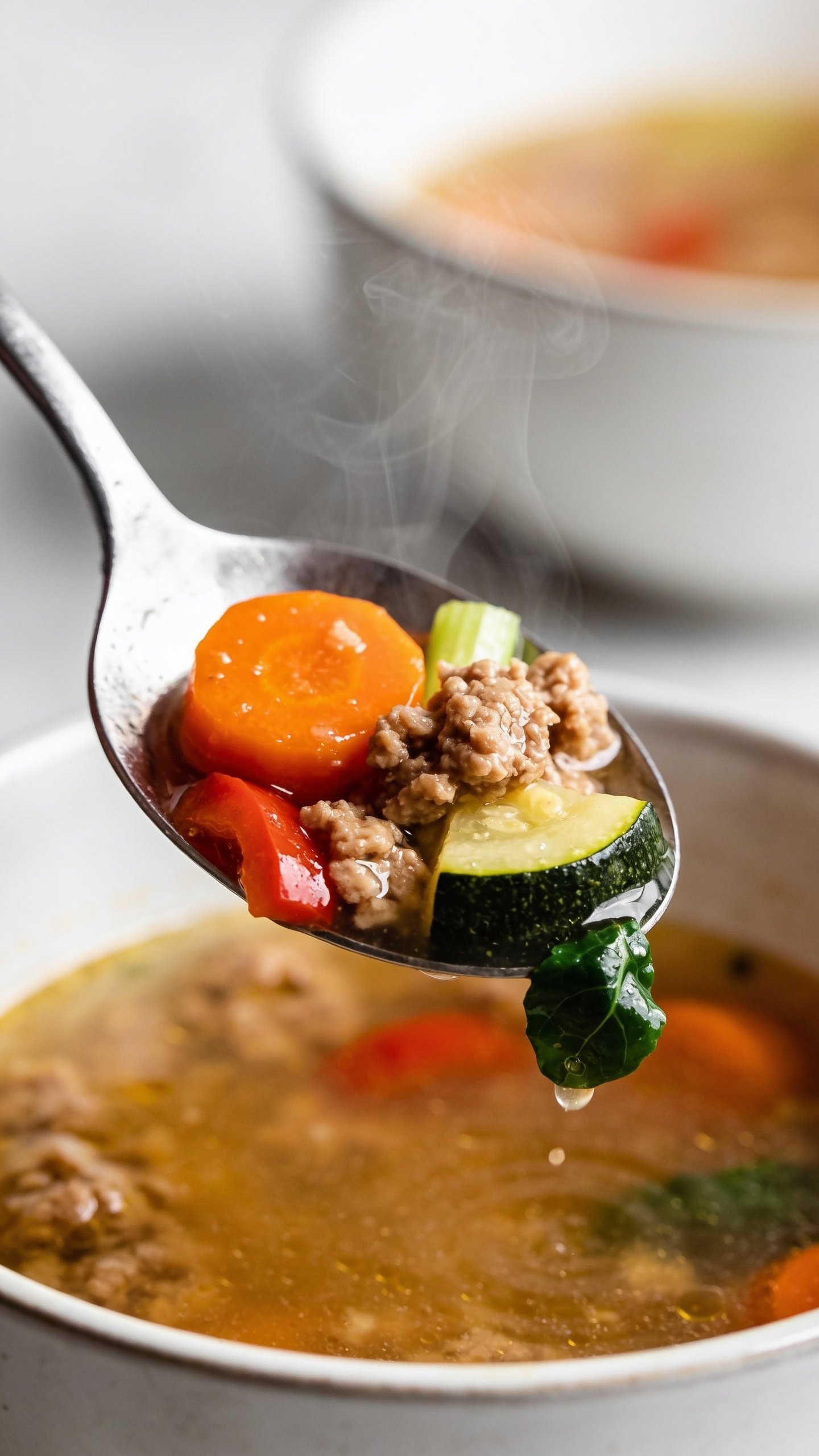 Dynamic action close-up of the same soup one second before a bite: a stainless soup spoon lifting the perfect mouthful from the bowl, hovering above the surface. The spoon holds glossy broth, a tender crumble of browned turkey, a coin of carrot, a piece of zucchini, a bit of red bell pepper, and a sliver of celery, with a tiny spinach leaf clinging to the edge; visible micro-droplets glistening. Gentle steam rising, background bowl blurred but clearly the same soup. Bright natural side light, shallow depth of field, 90–95% frame filled by the spoon and bowl, no hands or faces, premium magazine-style anticipation.