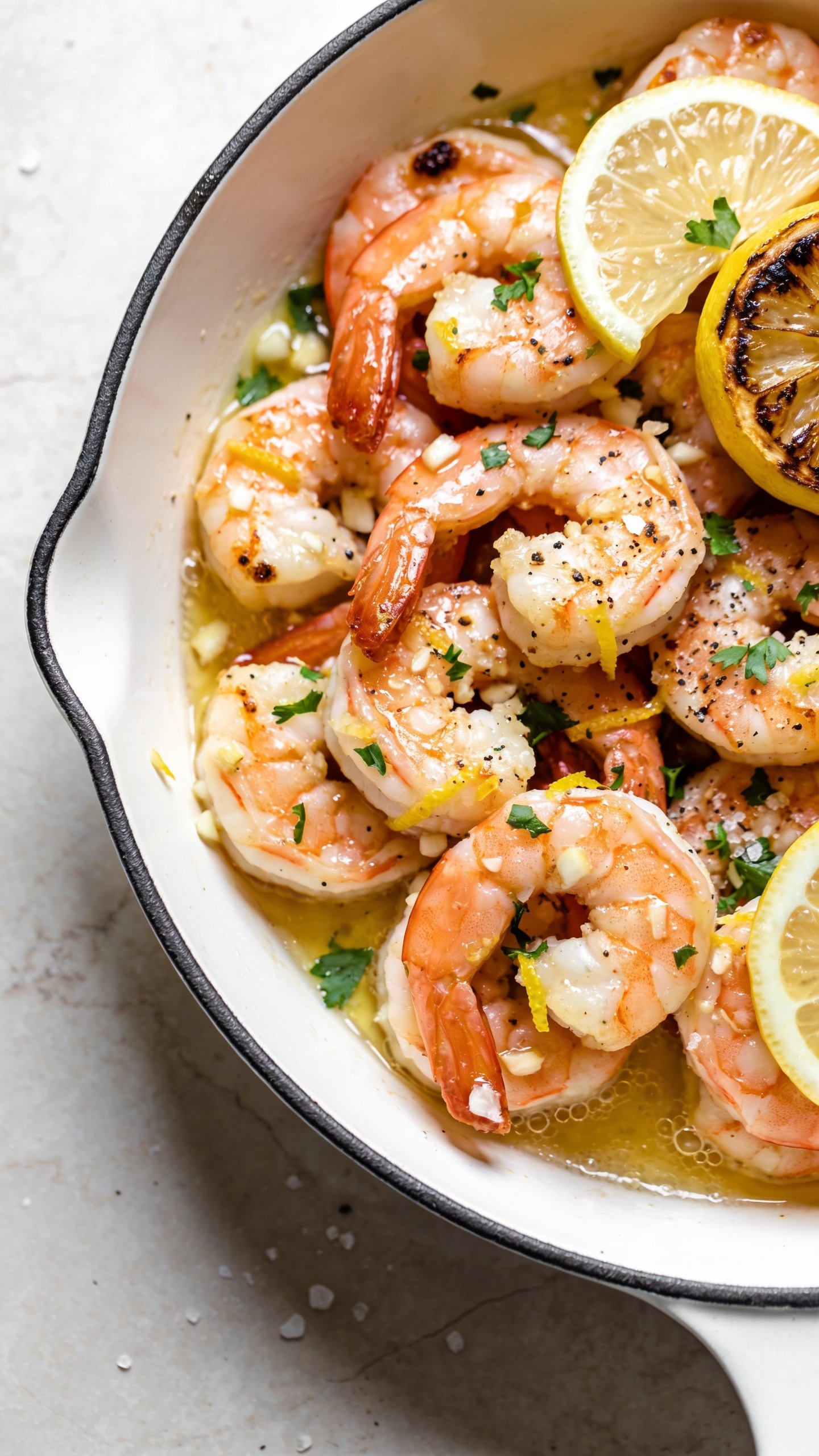 Ultra-tight overhead-to-45° angle close-up of the same finished lemon garlic shrimp piled in a shallow rimmed white skillet or platter: shrimp arranged in overlapping curls, glossy garlic-lemon butter pooling lightly in the crevices, specks of lemon zest and minced garlic clinging to the surfaces, sprinkled with fresh chopped parsley and a final grind of black pepper. Thin lemon slices and one lightly charred half-lemon add brightness. Capture crisp caramelized spots on shrimp edges, micro-bubbles in the sauce, and a few coarse sea salt flakes. Bright natural lighting, shallow depth of field focusing on the center cluster, food filling 90–95% of the frame, clean premium styling on a pale stone surface.