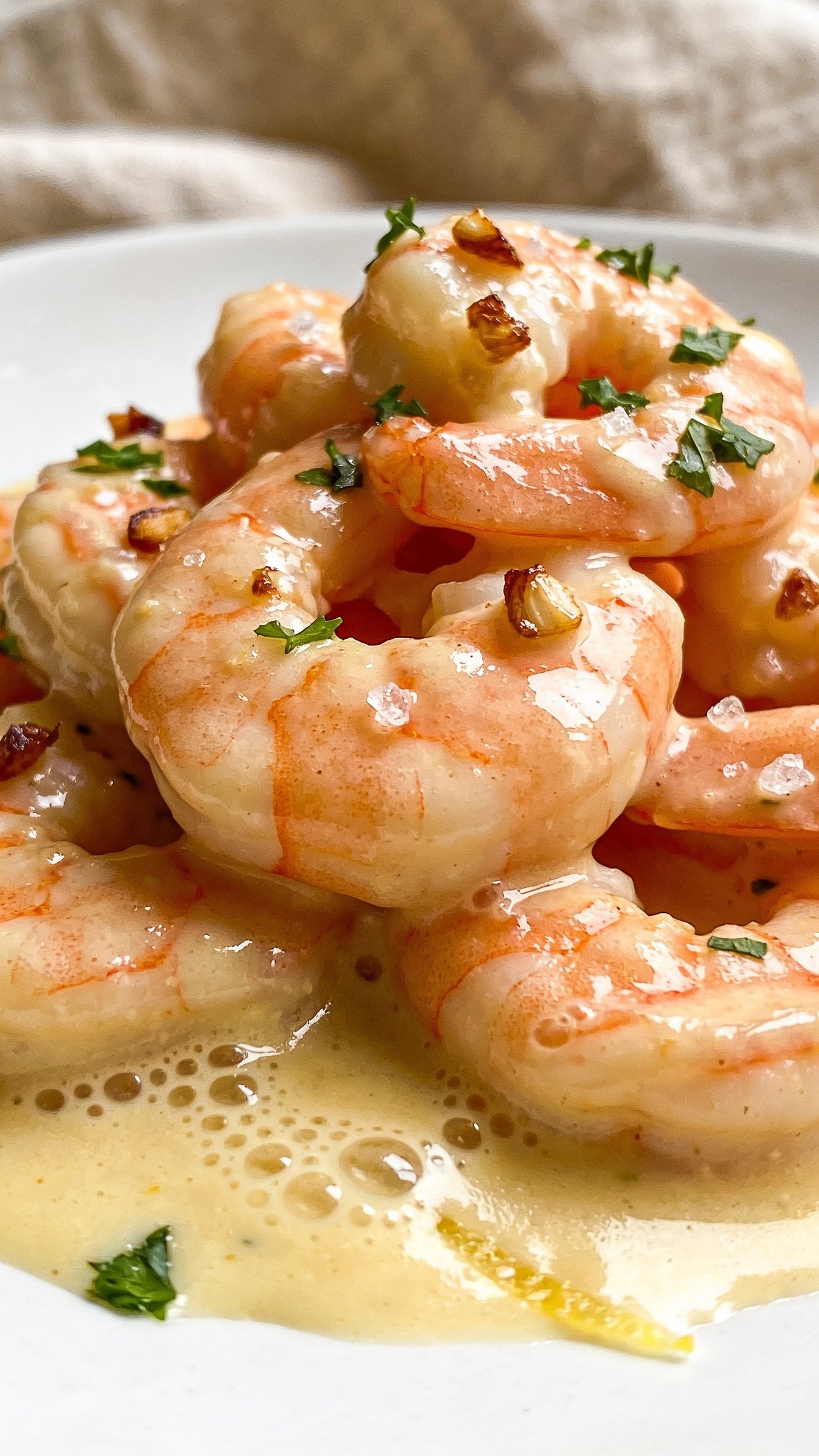 Extreme close-up angle from slightly overhead: a tight cluster of shrimp bathed in the luscious garlic cream, sauce glistening with micro-bubbles and buttery sheen, tiny browned garlic bits and chopped parsley scattered on top; a few coarse sea salt crystals catching light; faint lemon zest whisper on the surface; the edge of the white plate barely visible, warm linen blur in background; bright natural lighting, shallow depth of field isolating textures, food dominating 90â95% of the frame, same styling and color palette as the other shots.