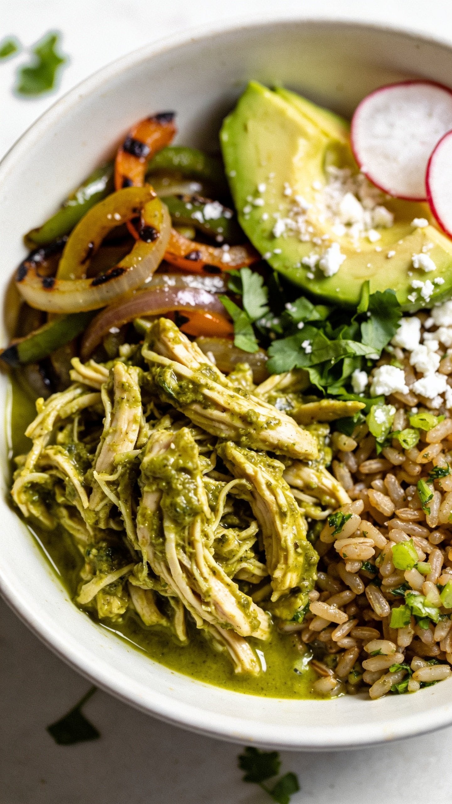 Beautiful close-up from a slightly overhead angle focusing on textures: the same finished bowl tightly framed so the saucy shredded salsa verde chicken and toppings dominate. Emphasize glossy salsa pooling lightly around the chicken, fluffy cilantro-lime brown rice grains, charred edges on roasted peppers and onions, creamy avocado with fine salt crystals, specks of cotija, fresh chopped cilantro scattered, and thin radish slices adding crisp contrast. Bright natural light, shallow depth of field, 90–95% food in frame, vibrant greens and warm char tones, premium food-blog styling, no utensils, no text.
