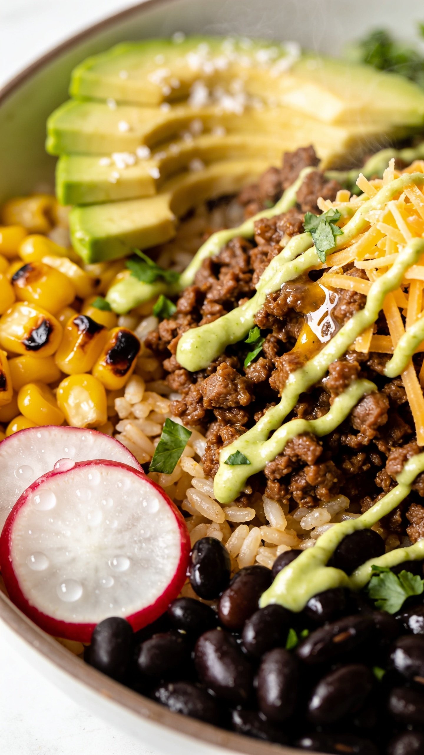 Ultra-close macro from a different angle (three-quarter overhead) of the same finished bowl emphasizing textures: glistening taco beef crumbles nestled over the rice blend, glossy black beans, crisp radish slices with tiny water droplets, charred corn with caramelized edges, avocado fan with flaky sea salt, fine shreds of cheddar lightly melting into the warm beef, scattered cilantro, and a delicate zigzag of lime crema. Vibrant colors, shallow depth of field with the beef and crema in sharp focus, bright natural light, subtle steam, clean minimal surface, 90–95% frame filled with food, premium food-blog styling, no text or hands.